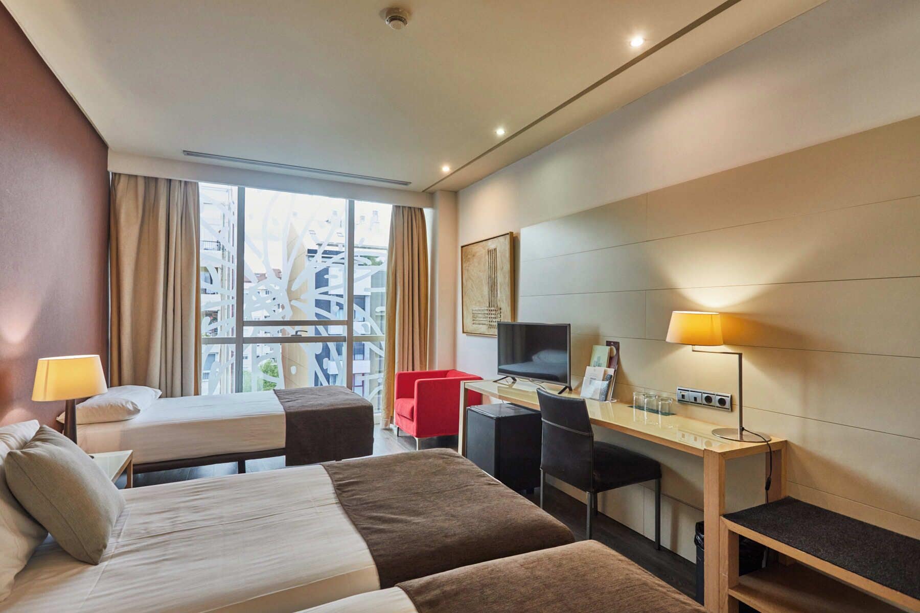 A hotel room with two beds, a desk, a chair, a TV, and a window with curtains.