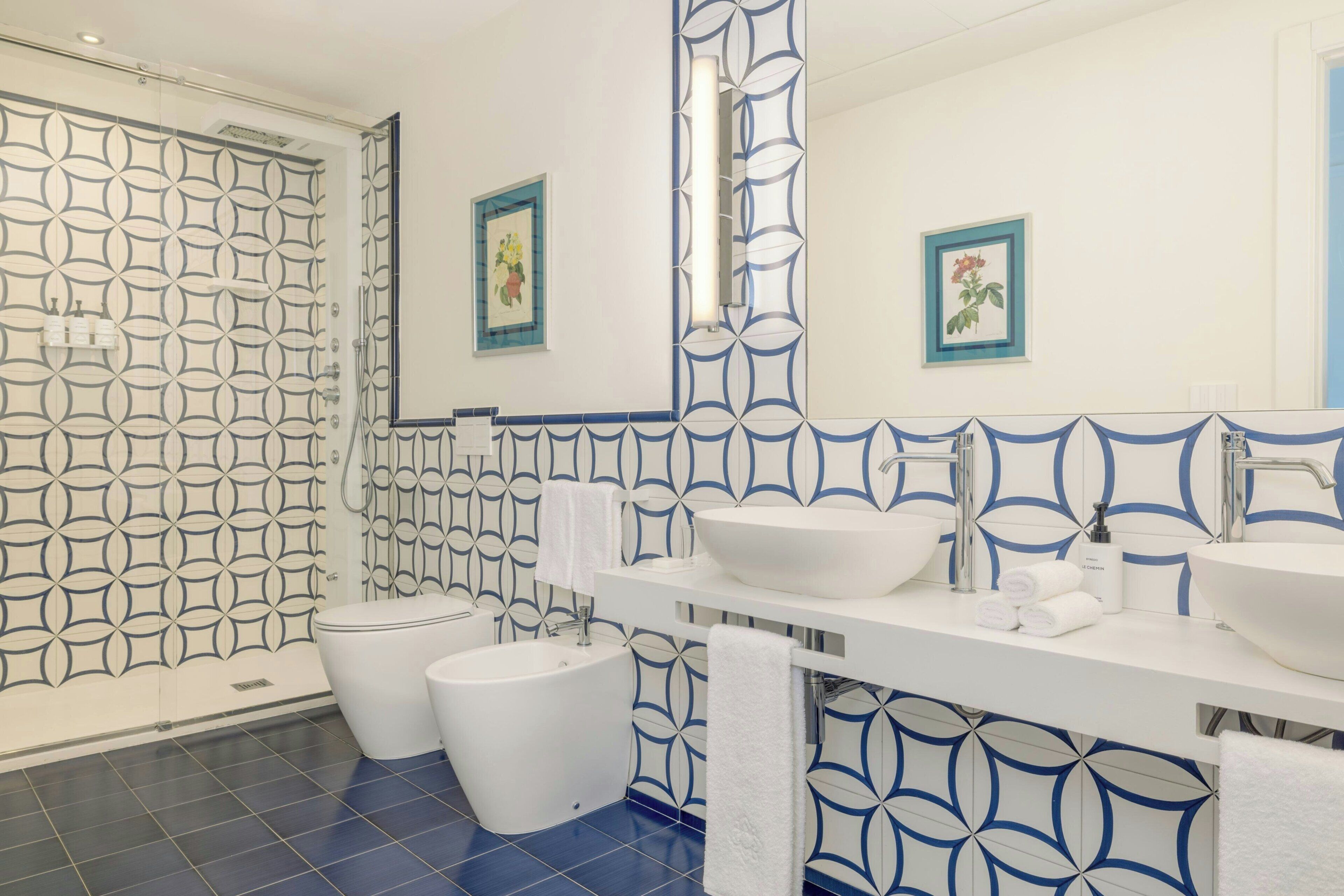 A modern bathroom with a geometric tile pattern, a large mirror, two sinks, and a shower.