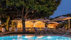 Un área de piscina con sillones de descanso y sombrillas, un bar con asientos y un árbol grande que proporciona sombra.