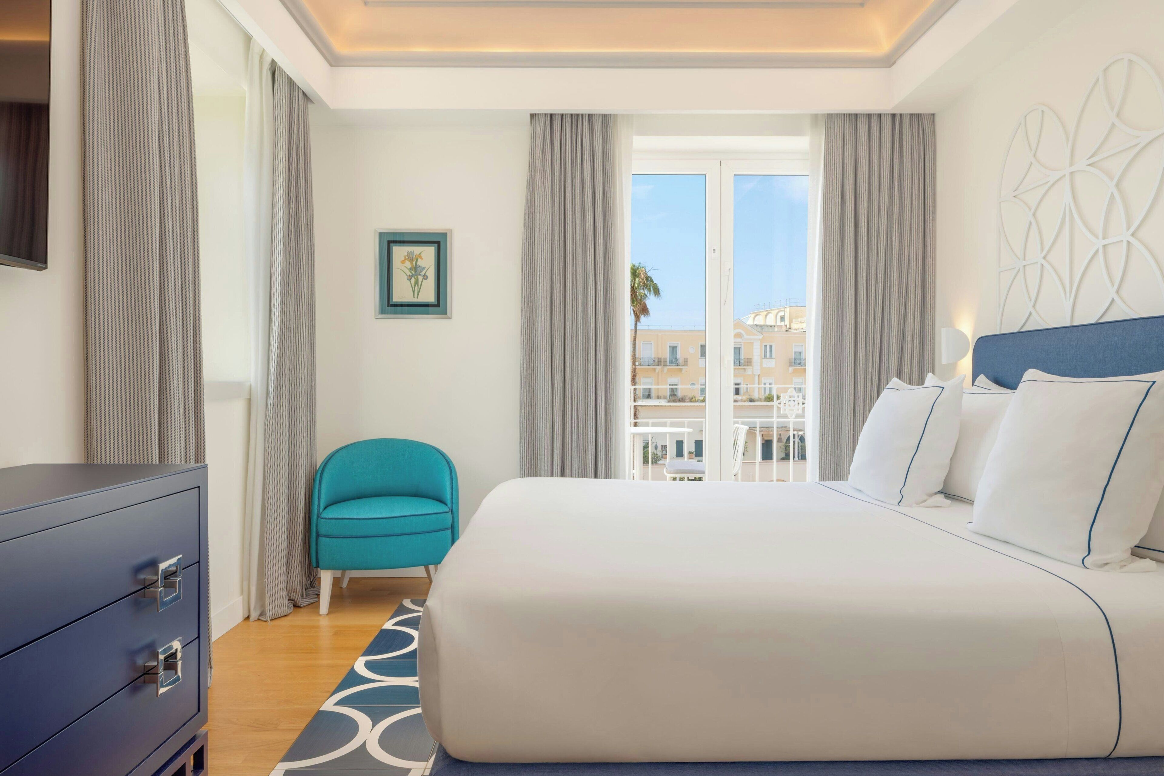 A hotel room with a bed, a blue chair, a dresser, and a view of buildings through the window.