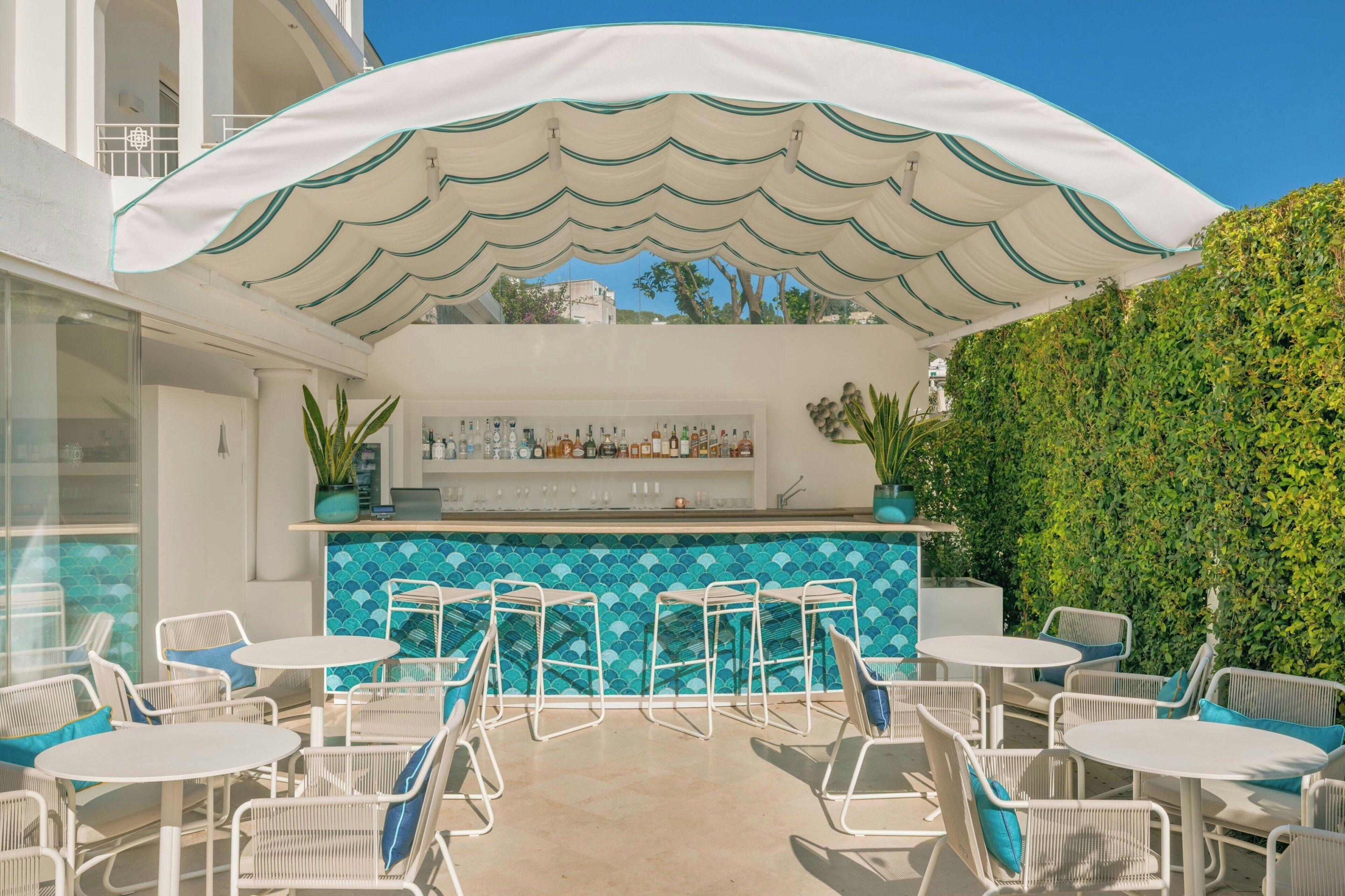 Un área de bar al aire libre con una sombrilla blanca, sillas blancas y una pared de piscina con azulejos azules.