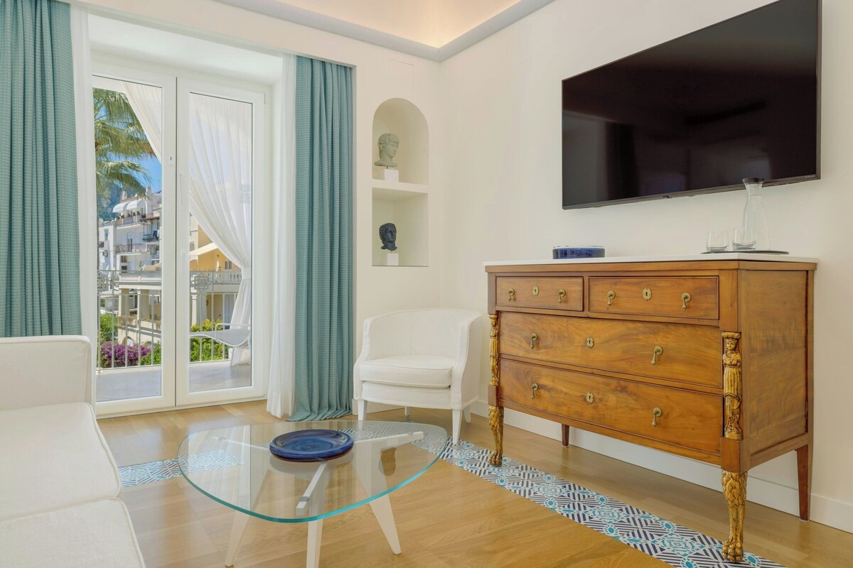 Een woonkamer met een glazen tafel, een witte bank, een houten kast en een aan de muur bevestigde televisie.