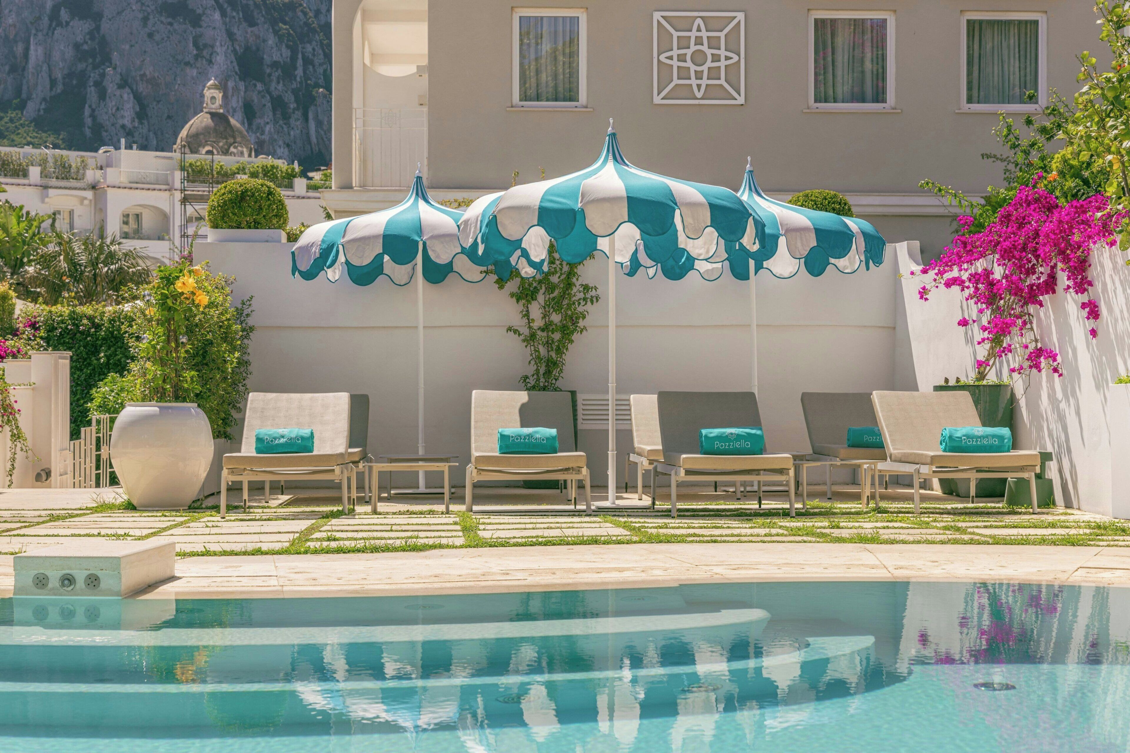 Un área junto a la piscina con sillones de descanso y sombrillas, una piscina cristalina y un edificio con cúpula al fondo.