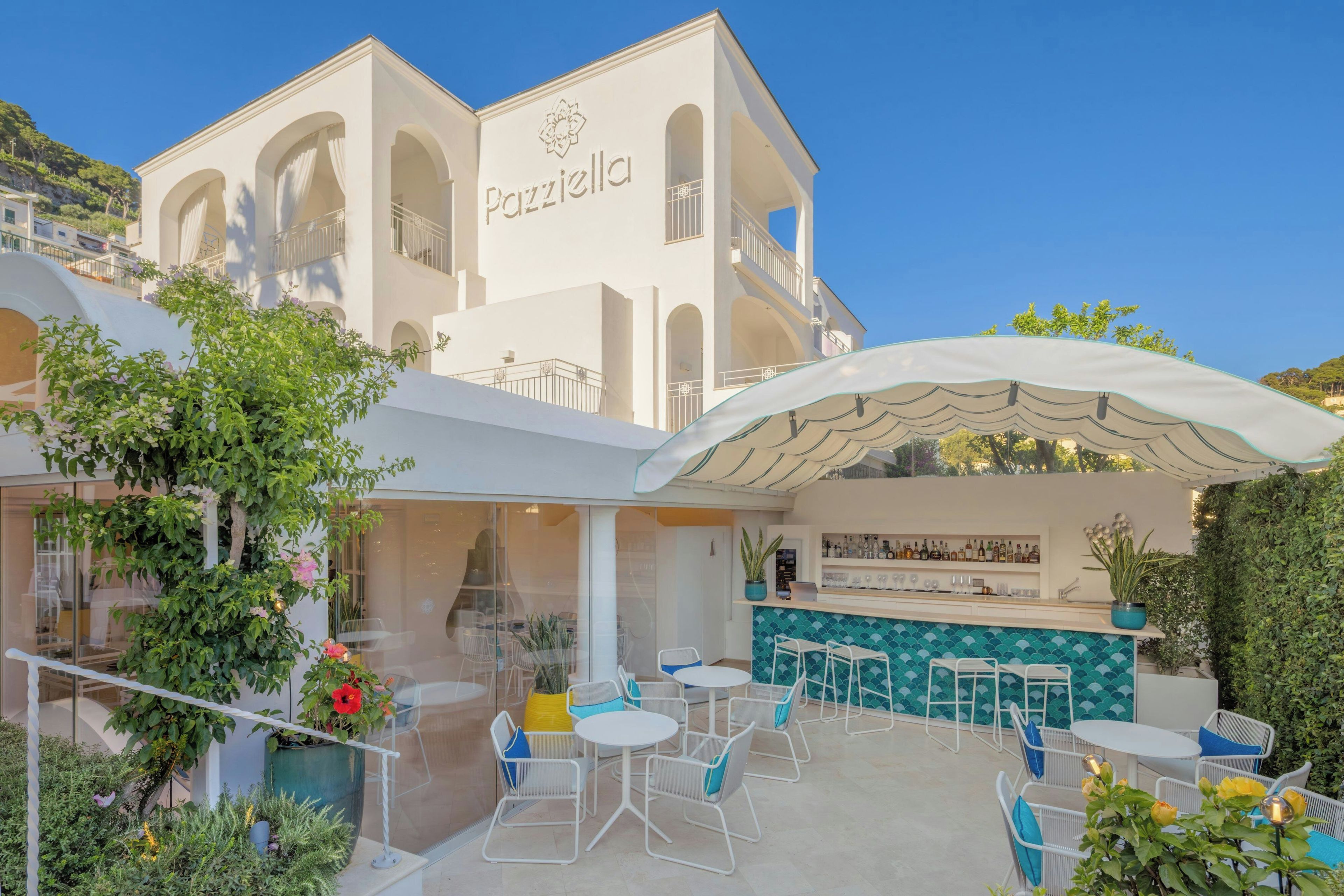 Un edificio con el nombre 'Pazzella' en la fachada, con terraza y mesas y sillas, y una zona de bar con salpicadero de azulejos.