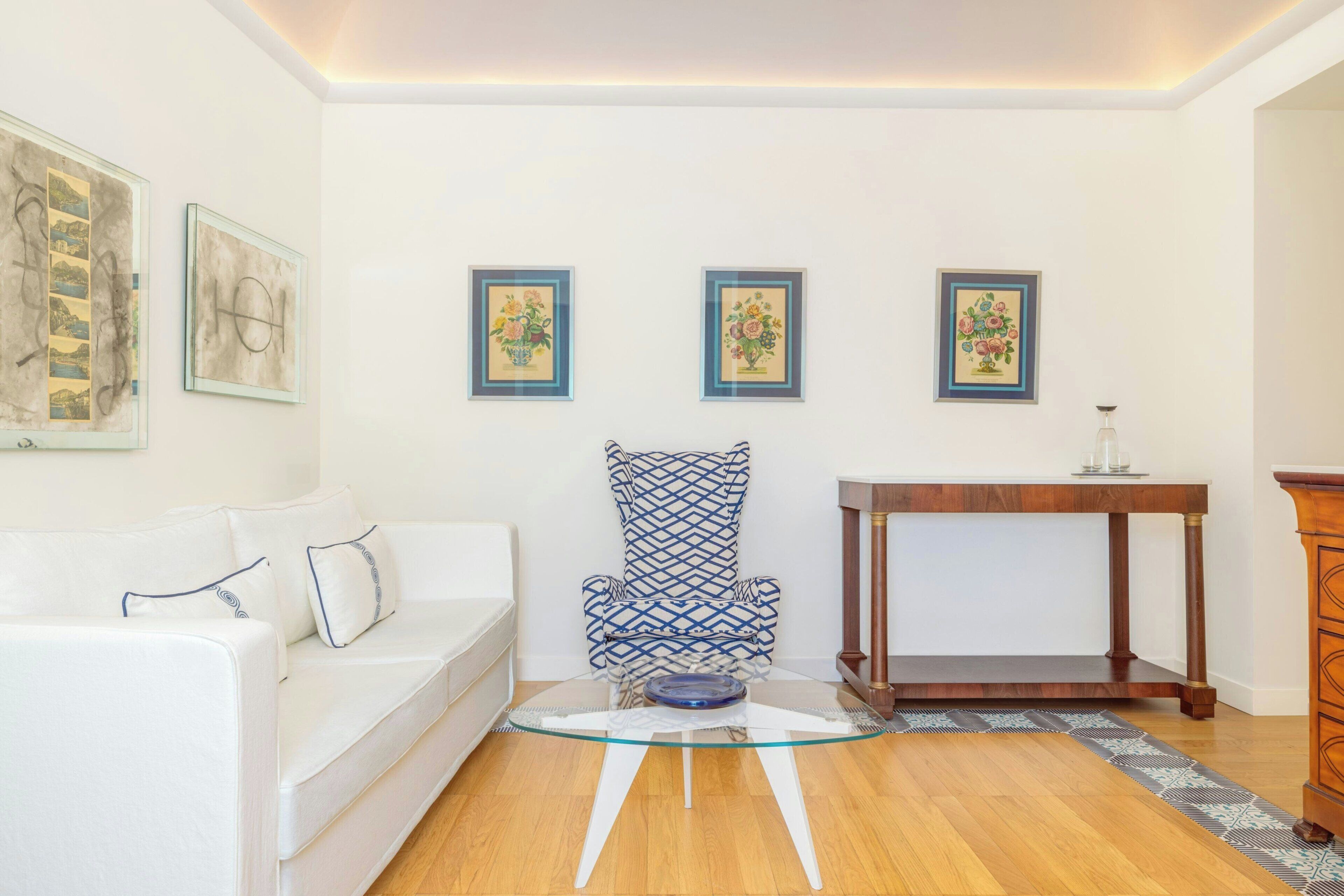 Una sala de estar con un sofá blanco, un sillón estampado, una mesa de centro de vidrio y una mesita de noche de madera.