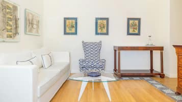 Una sala de estar con un sofá blanco, un sillón estampado, una mesa de centro de vidrio y una mesita de noche de madera.
