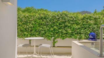 A balcony with a white table and two chairs, a blue decorative object, and a green hedge wall.