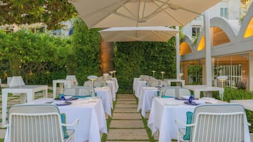 An outdoor dining area with white tables and chairs, a covered walkway, and a building with arched windows in the background.
