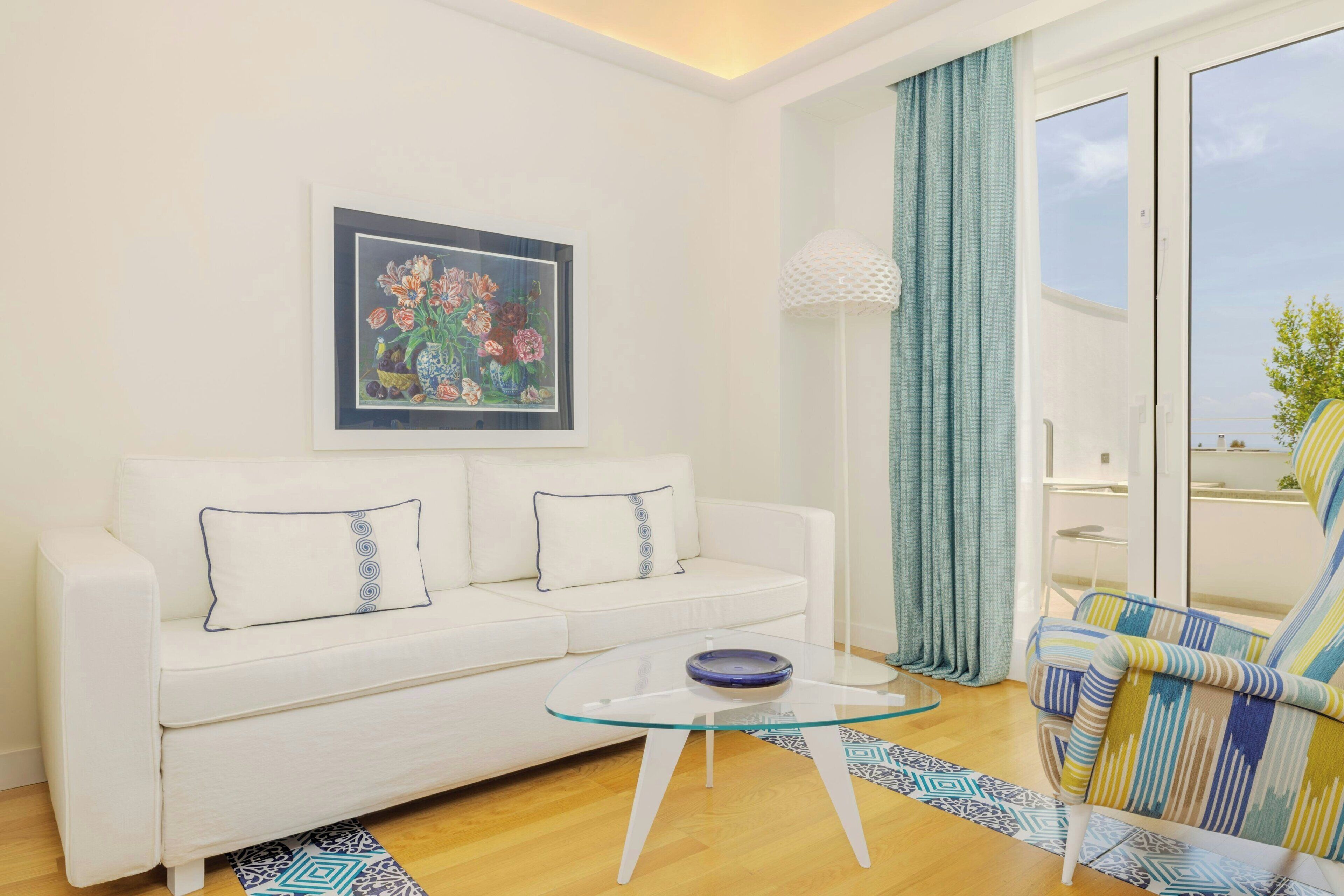 Una sala de estar con un sofá blanco, una mesa de centro de cristal, un sillón colorido, una obra de arte floral enmarco y vistas al exterior a través de una ventana.