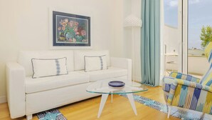 Una sala de estar con un sofá blanco, una mesa de centro de cristal, un sillón colorido, una obra de arte floral enmarco y vistas al exterior a través de una ventana.