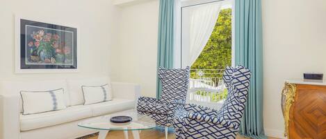 Una sala de estar moderna con un sofá blanco, un sillón estampado, una mesa de centro de vidrio y vistas a la vegetación a través de cortinas traslúcidas.
