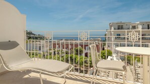 Un balcon avec deux fauteuils et une petite table, donnant sur une zone côtière avec des bâtiments et un ciel dégagé.