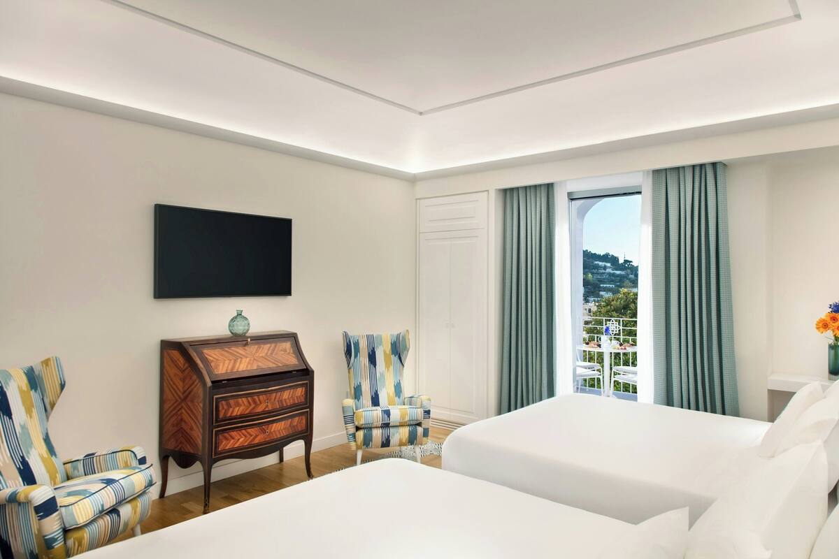 Une chambre d’hôtel avec deux lits, un téléviseur fixé au mur, une commode en bois et une vue sur l’extérieur par une fenêtre avec des rideaux.