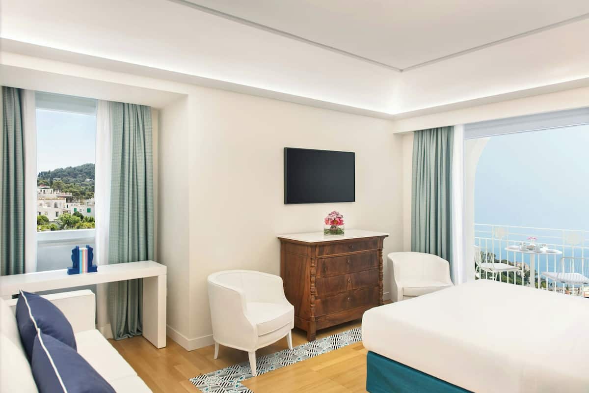 Une chambre d’hôtel moderne avec un lit, deux chaises blanches, une commode en bois, un téléviseur fixé au mur et une vue sur la mer.