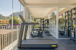 Un gimnasio al aire libre techado con equipo de ejercicios, incluyendo una cinta de correr y máquinas de pesas, con vistas a una zona residencial con casas y vegetación.