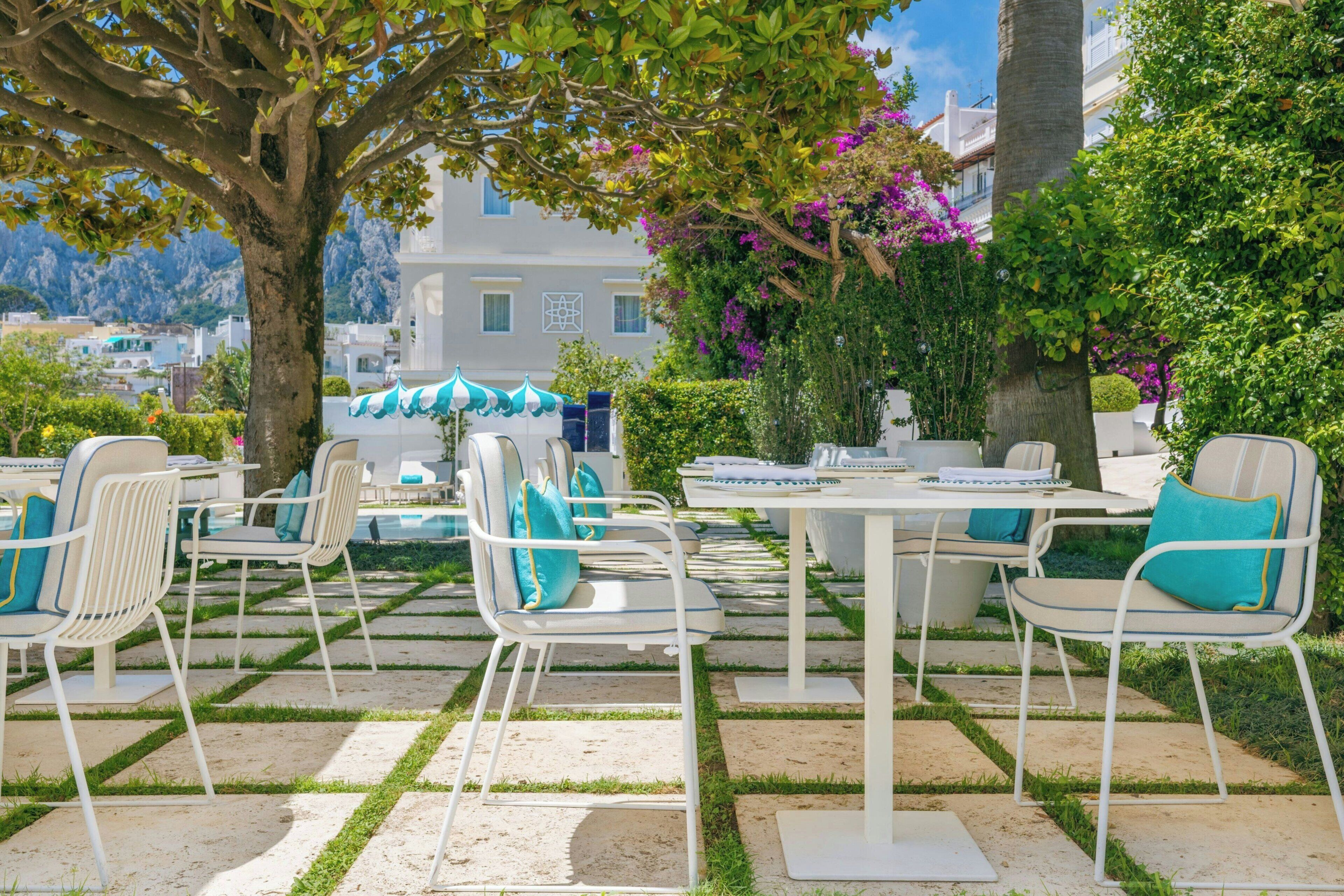 Een terras met witte metalen stoelen en tafels, omringd door groen en een uitzicht op gebouwen op de achtergrond.