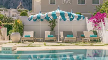 Área da piscina com espreguiçadeiras e guarda-sóis, um vaso decorativo grande e um edifício com cúpula ao fundo.