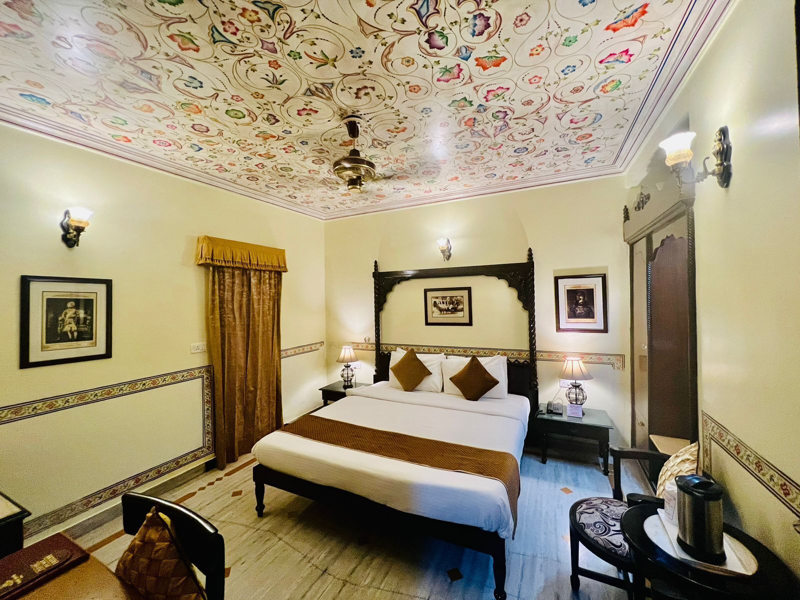 A hotel room with a large bed, a ceiling with floral patterns, a wooden headboard, and framed pictures on the walls.