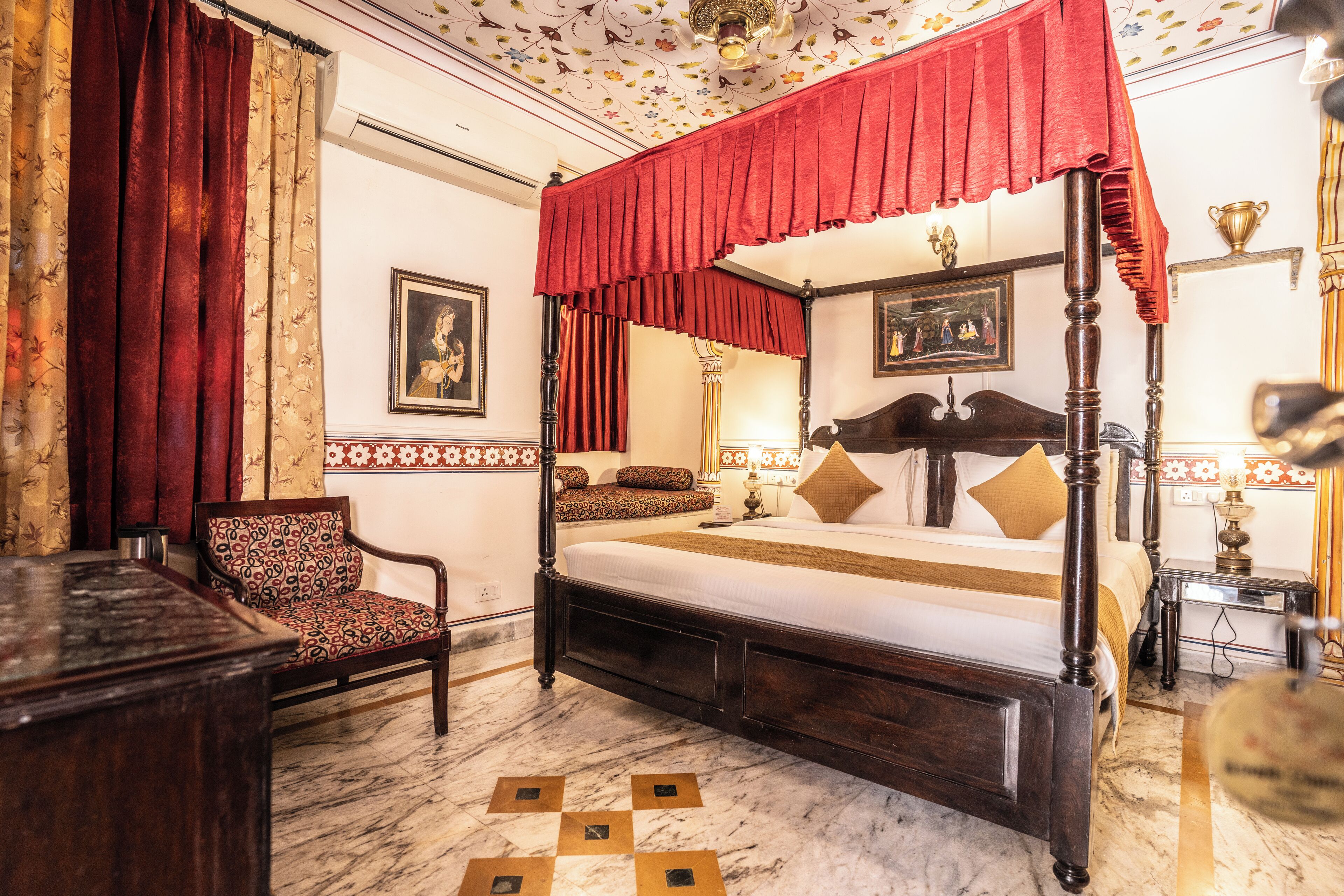 A four-poster canopy bed with a red canopy, a wooden chair with a patterned cushion, and a marble floor with geometric patterns.