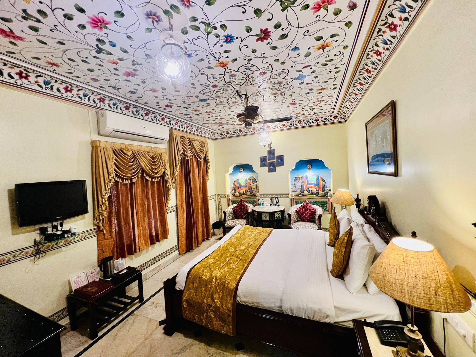 A hotel room with a bed, a television, a desk, and a ceiling with floral patterns.