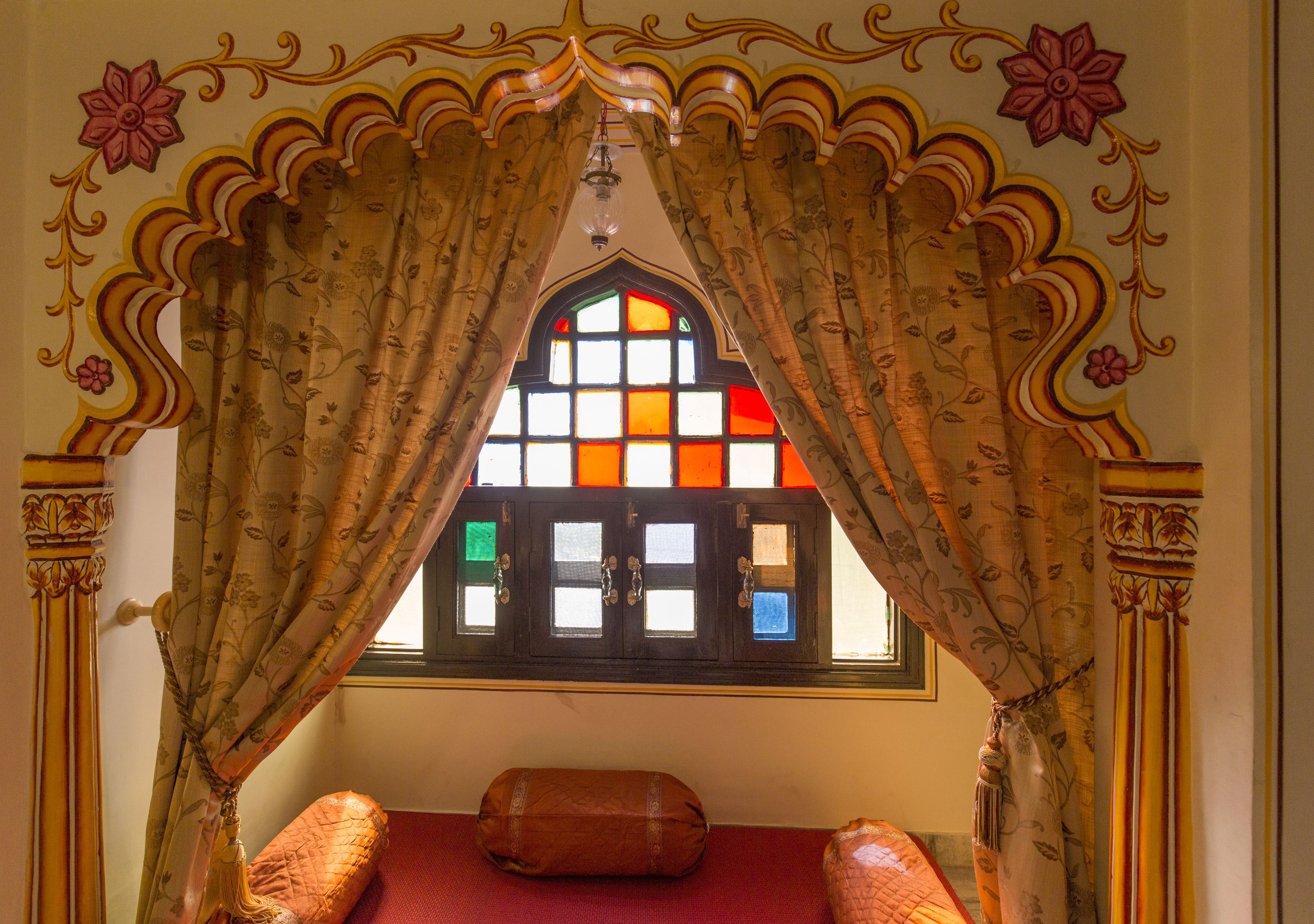 Una habitación con ventana enmarcada por cortinas ornamentadas, alfombra roja y almohada decorativa.