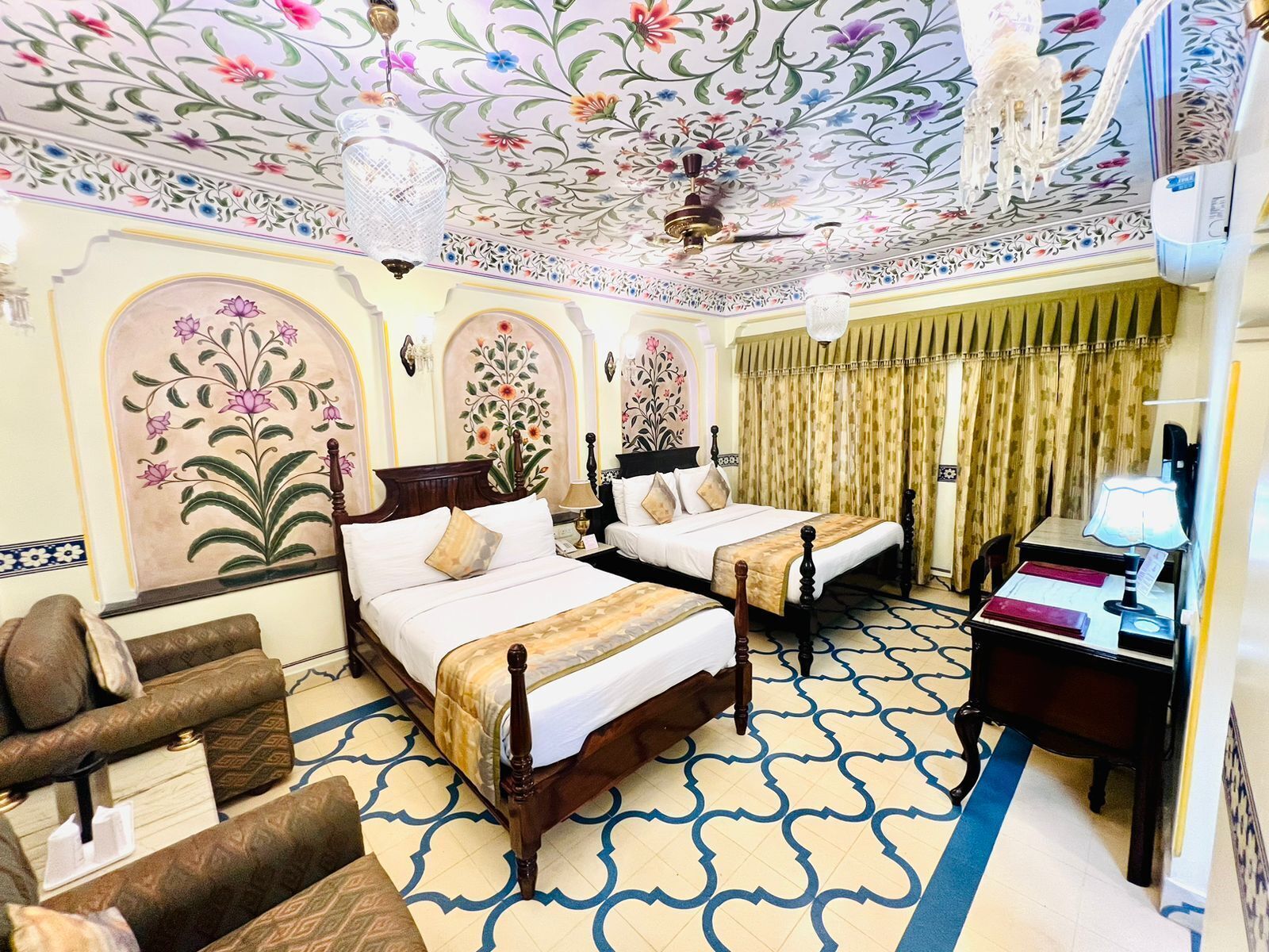 A hotel room with two beds, a desk, a chair, and a ceiling with floral patterns.