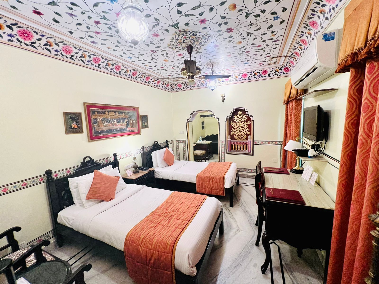 A hotel room with two beds, a desk, a chair, a mirror, and a ceiling with floral patterns.