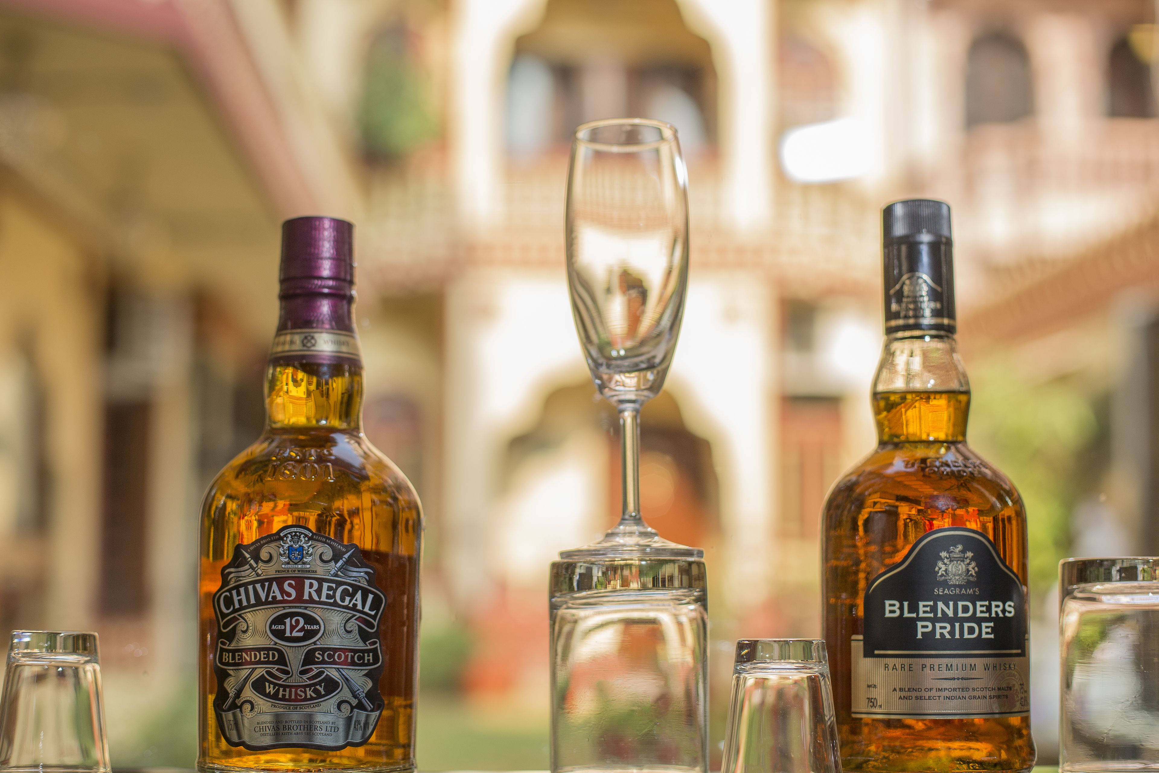 Two bottles of whiskey and a wine glass on a table with a blurred building in the background.