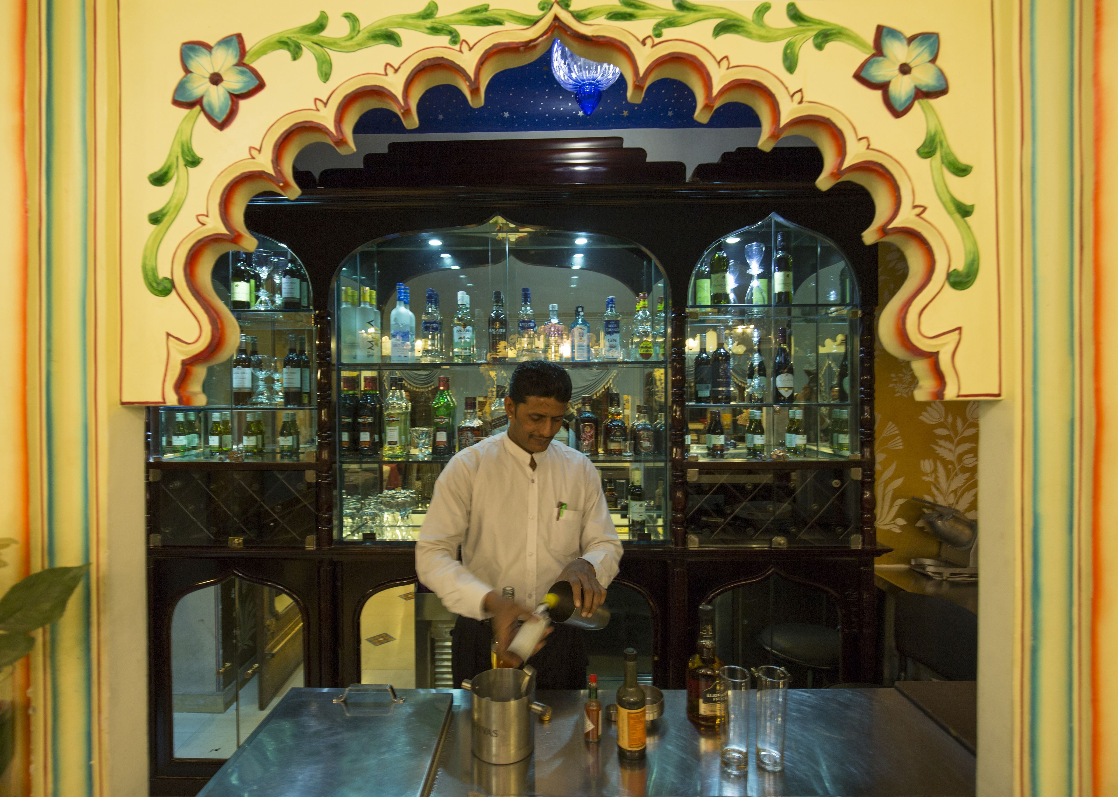 A bar area with a bartender preparing drinks. There are shelves with various bottles and glasses behind the bartender.