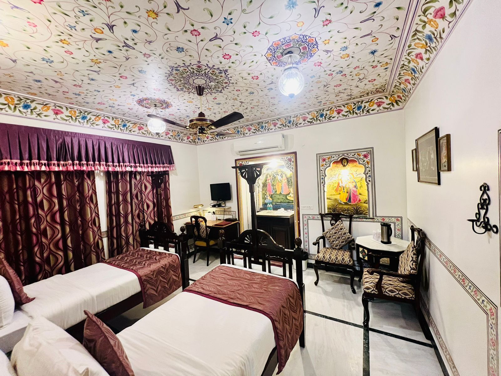 Habitación de hotel con dos camas, un escritorio, una silla, un televisor y un techo con diseños florales.