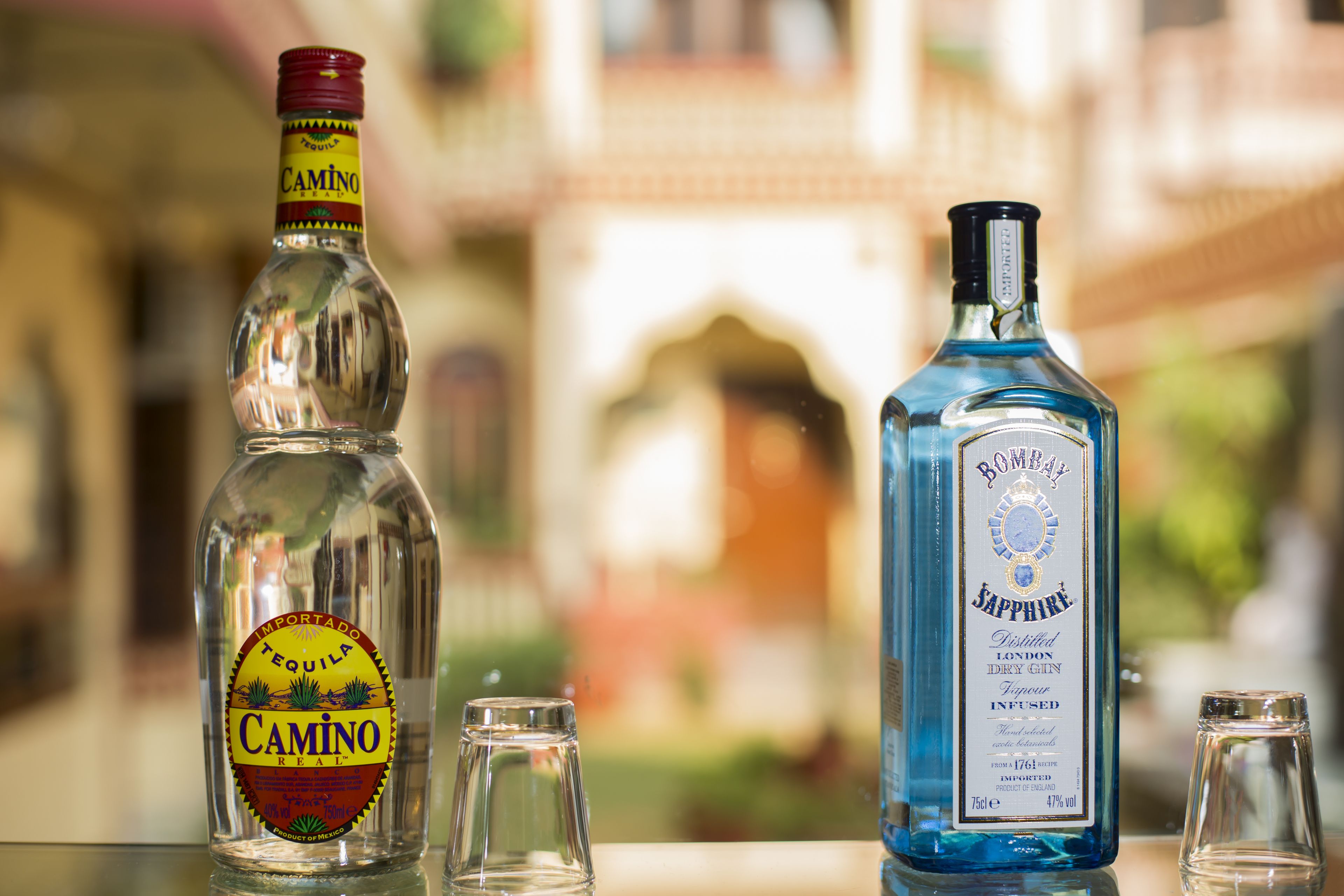 Zwei alkoholische Flaschen, eine mit dem Aufdruck 'Camino' und die andere 'Bombay Sapphire', mit einem unscharfen Gebäude im Hintergrund.