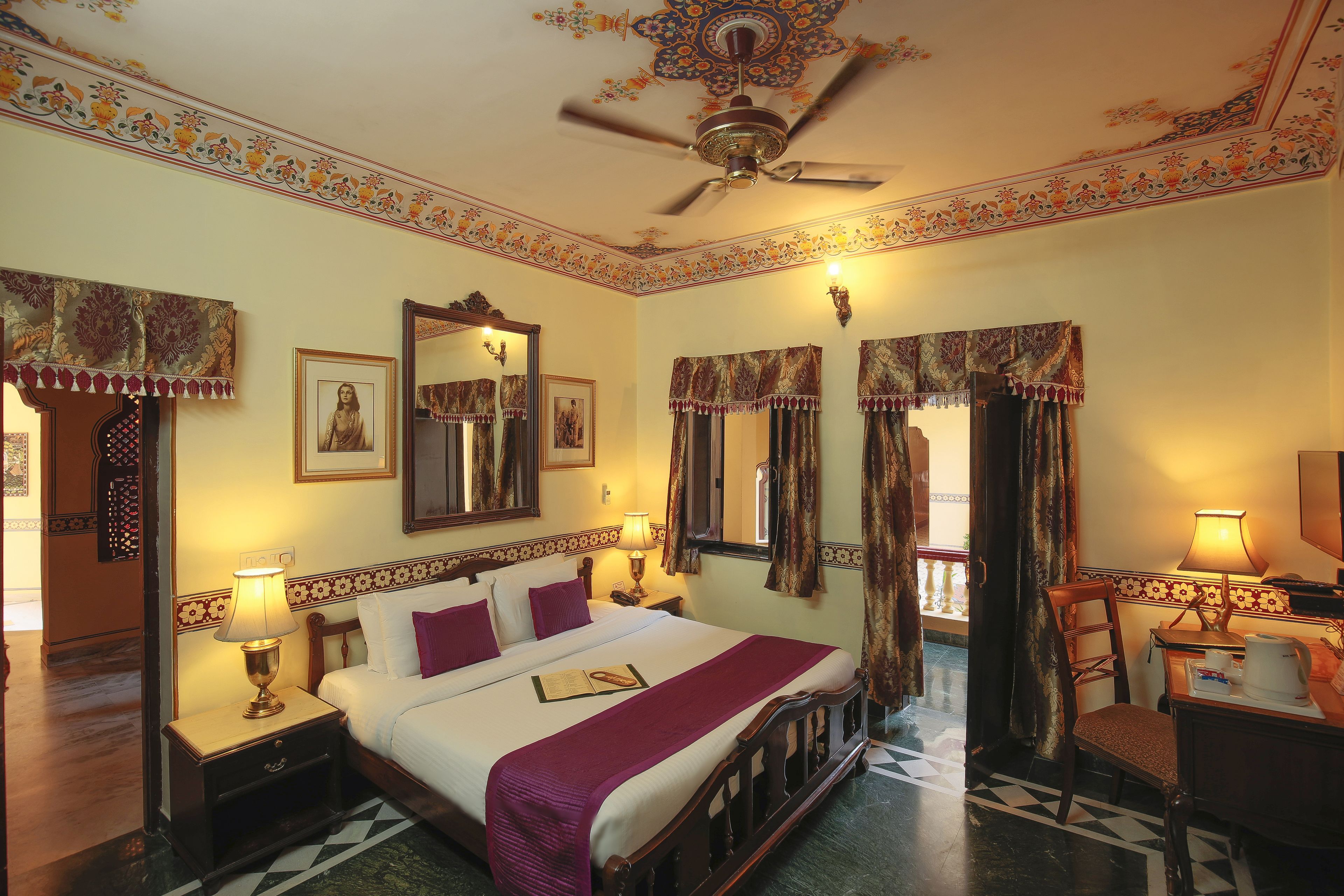 Habitación de hotel con una cama grande, ventilador de techo, decoraciones de pared ornamentales y un escritorio con lámpara.