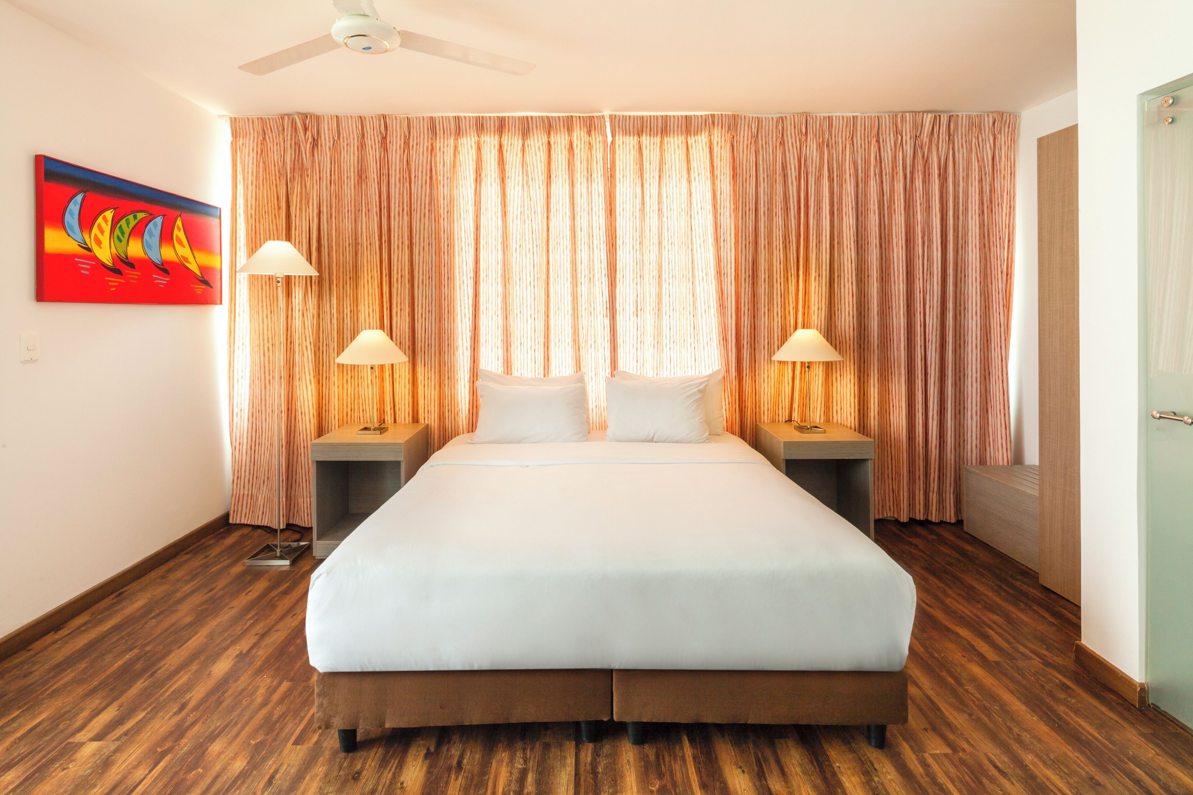 Habitación de hotel con una cama grande, dos mesitas de noche, un ventilador de techo y un cuadro en la pared.