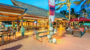 An outdoor dining area with wooden tables and chairs, a large umbrella, and a tiki statue.