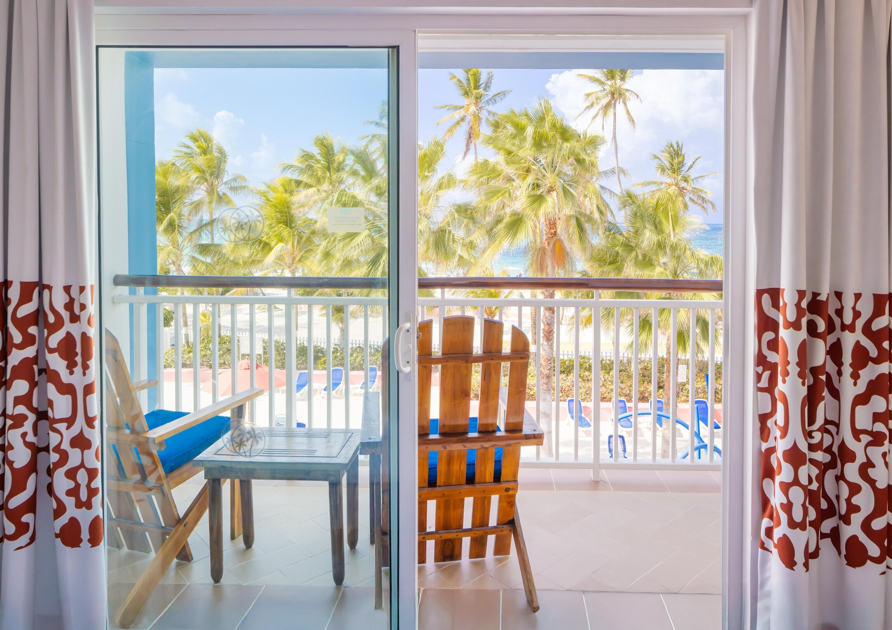 Una habitación con puerta de vidrio que da a un balcón con vista a palmeras y una piscina.