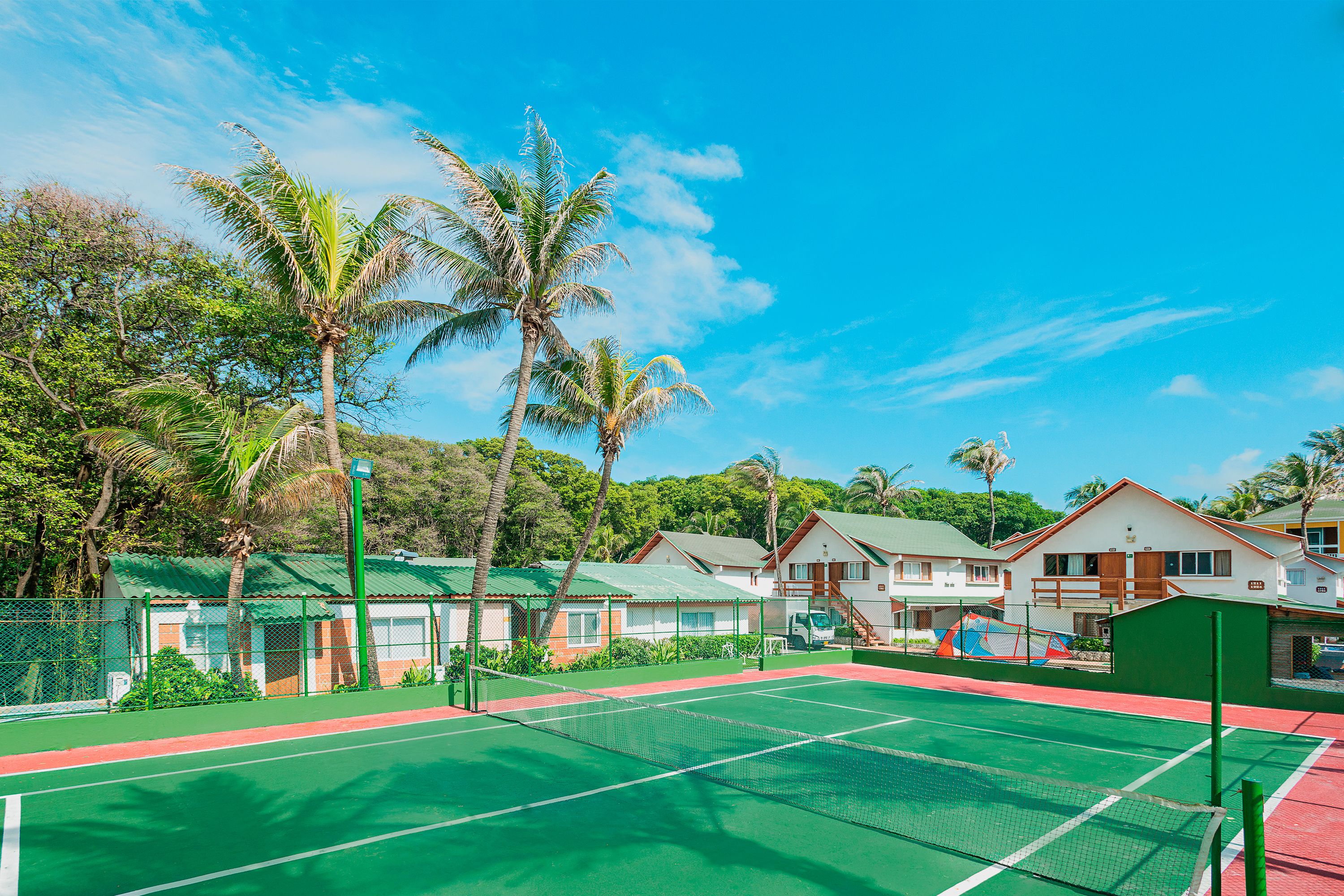 A tennis court with a net, surrounded by green fencing, palm trees, and houses with green roofs.