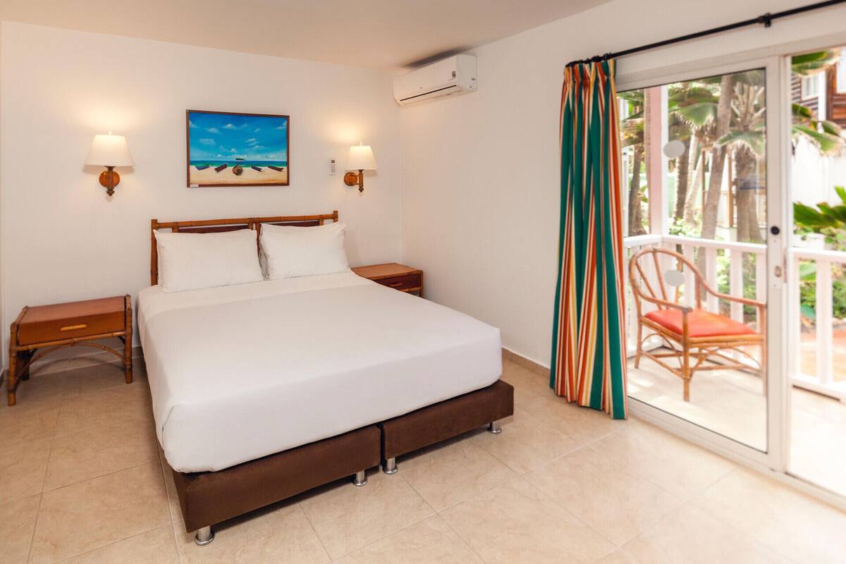 Ein Hotelzimmer mit einem Bett, zwei Nachttischen, einem Wandbild, einer Klimaanlage und einer Glastür, die zu einem Außenbereich mit einem Stuhl führt.