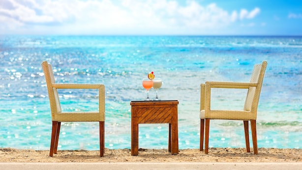 Zwei Korbstühle stehen vor einem Holztisch, auf dem zwei Getränke stehen. Im Hintergrund ein Strand mit klarem, blauem Wasser und einem strahlend blauen Himmel.