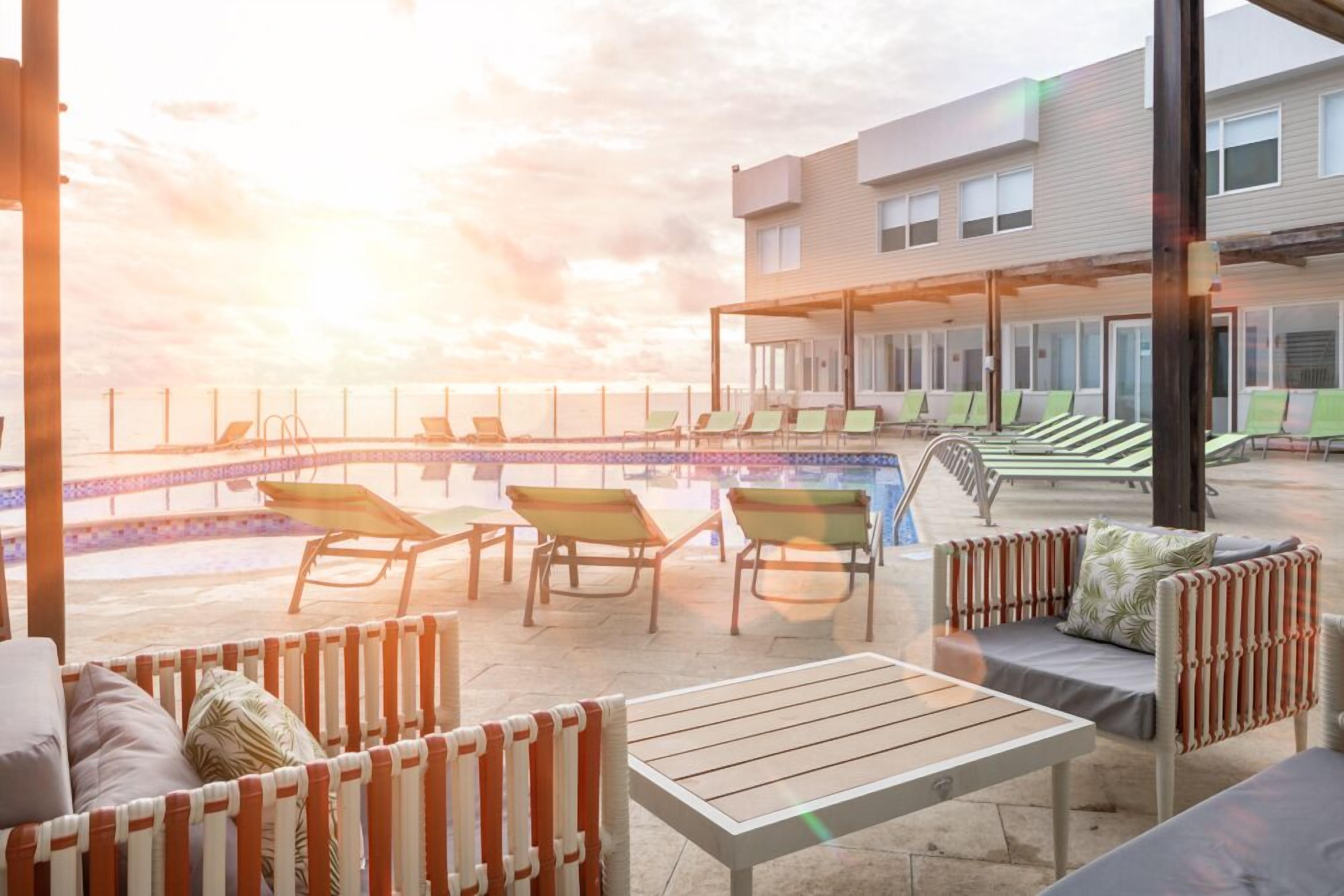 Uma área externa com cadeiras de descanso e uma mesa, com vista para uma piscina e um edifício com várias janelas.
