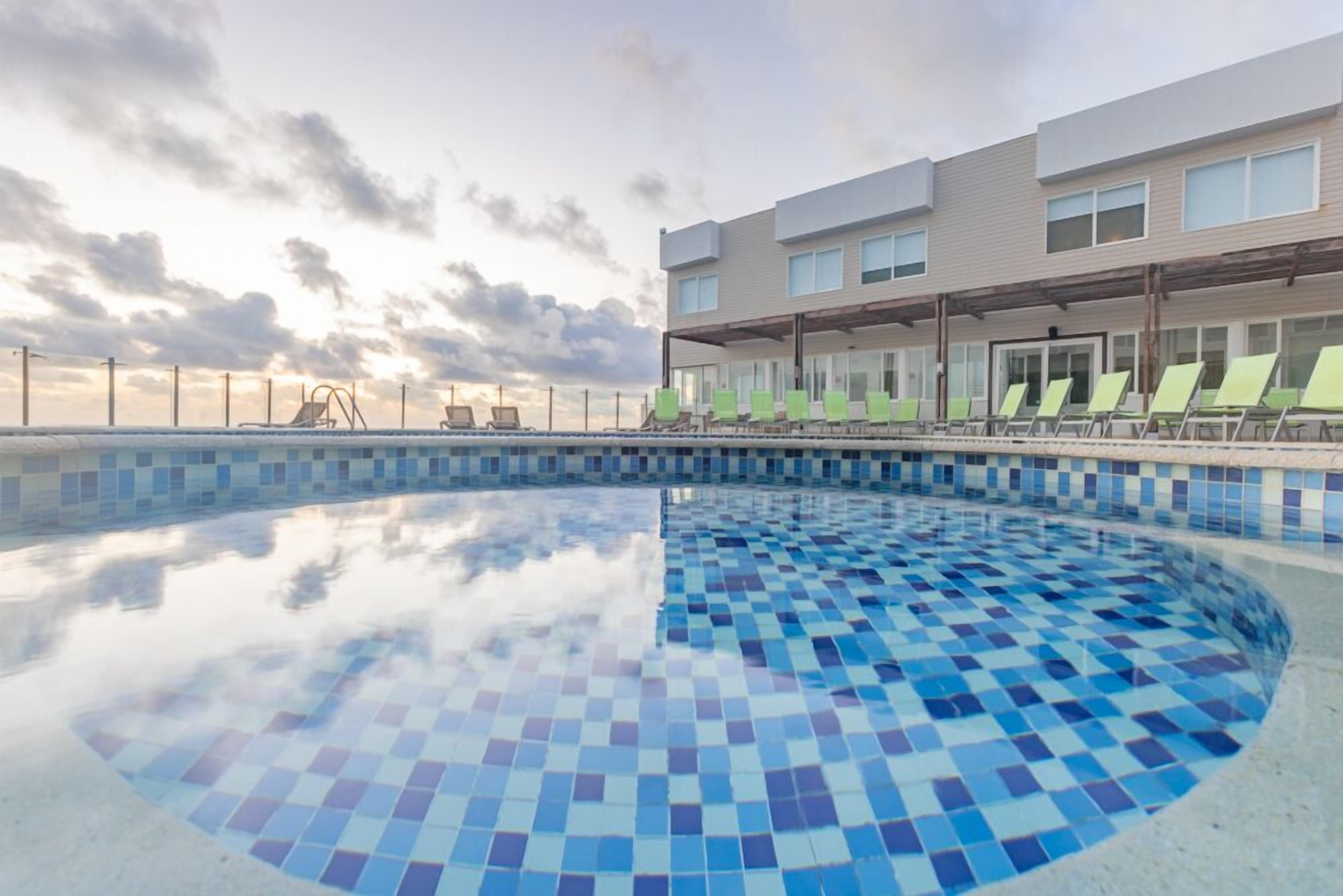 Um hotel moderno com piscina revestida em mosaicos azuis, área de estar coberta com cadeiras verdes e vista desobstruída para o pôr do sol.