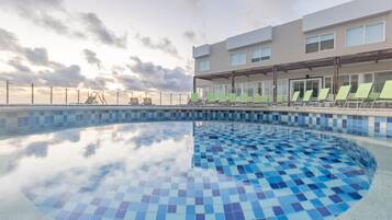 Um hotel moderno com piscina revestida em mosaicos azuis, área de estar coberta com cadeiras verdes e vista desobstruída para o pôr do sol.