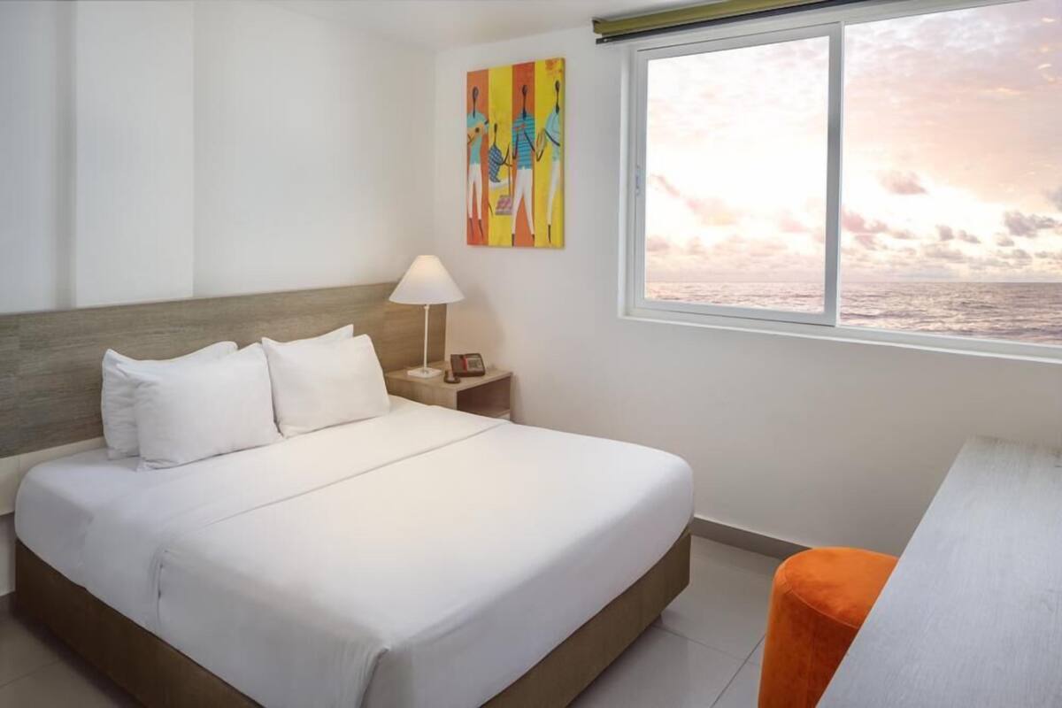 A hotel room with a bed, a nightstand, a lamp, a painting, and a window with a view of the sea.