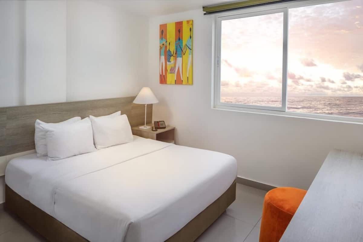 A hotel room with a bed, a nightstand, a lamp, a painting, and a window with a view of the sea.