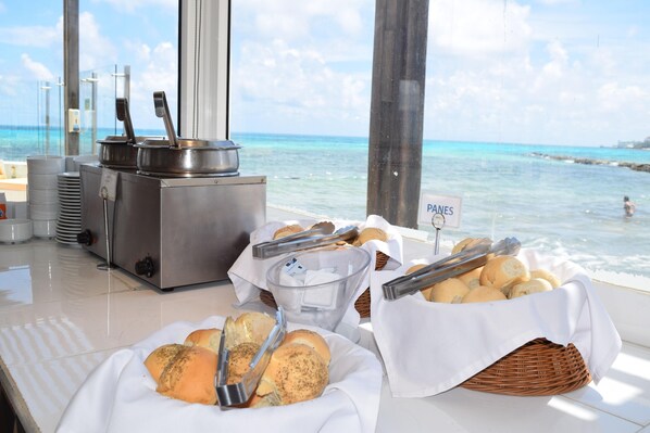 A buffet setup with bread, buns, and a sign indicating 'PANES' with a view of the ocean.