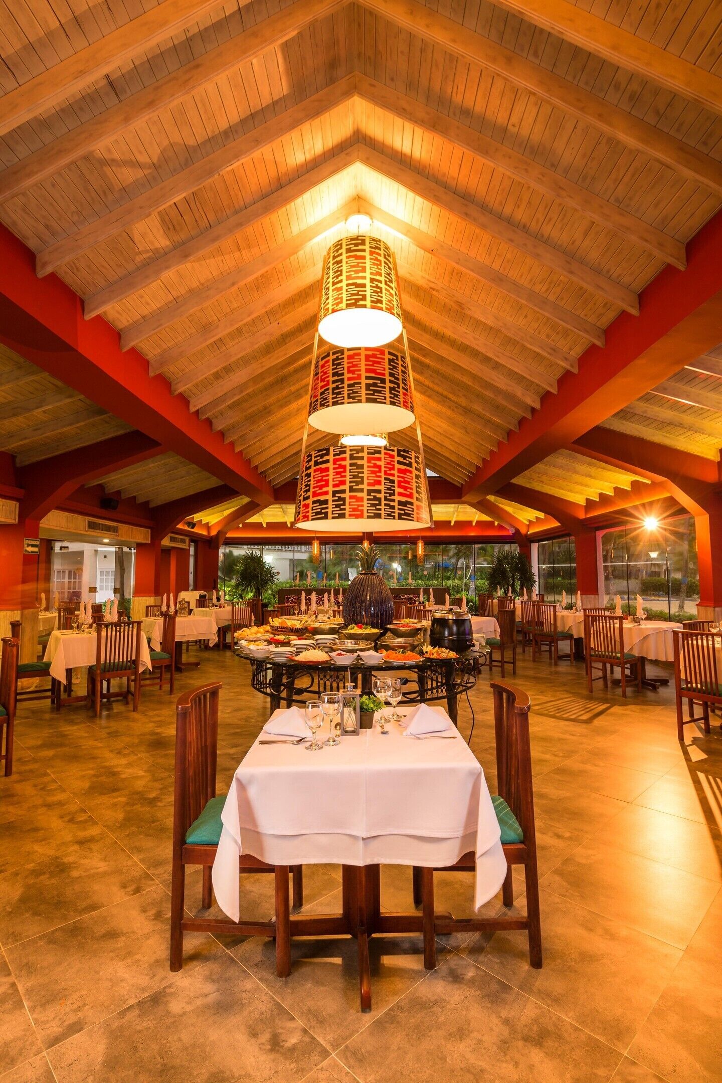 Un comedor con una mesa central dispuesta para una comida, rodeada de sillas, bajo un techo de madera con una lámpara decorativa.