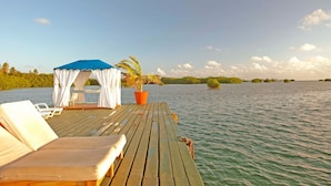 A wooden deck with lounge chairs and a canopy, overlooking a calm body of water with greenery in the background. - Decameron Marazul - All Inclusive (San Andrés)