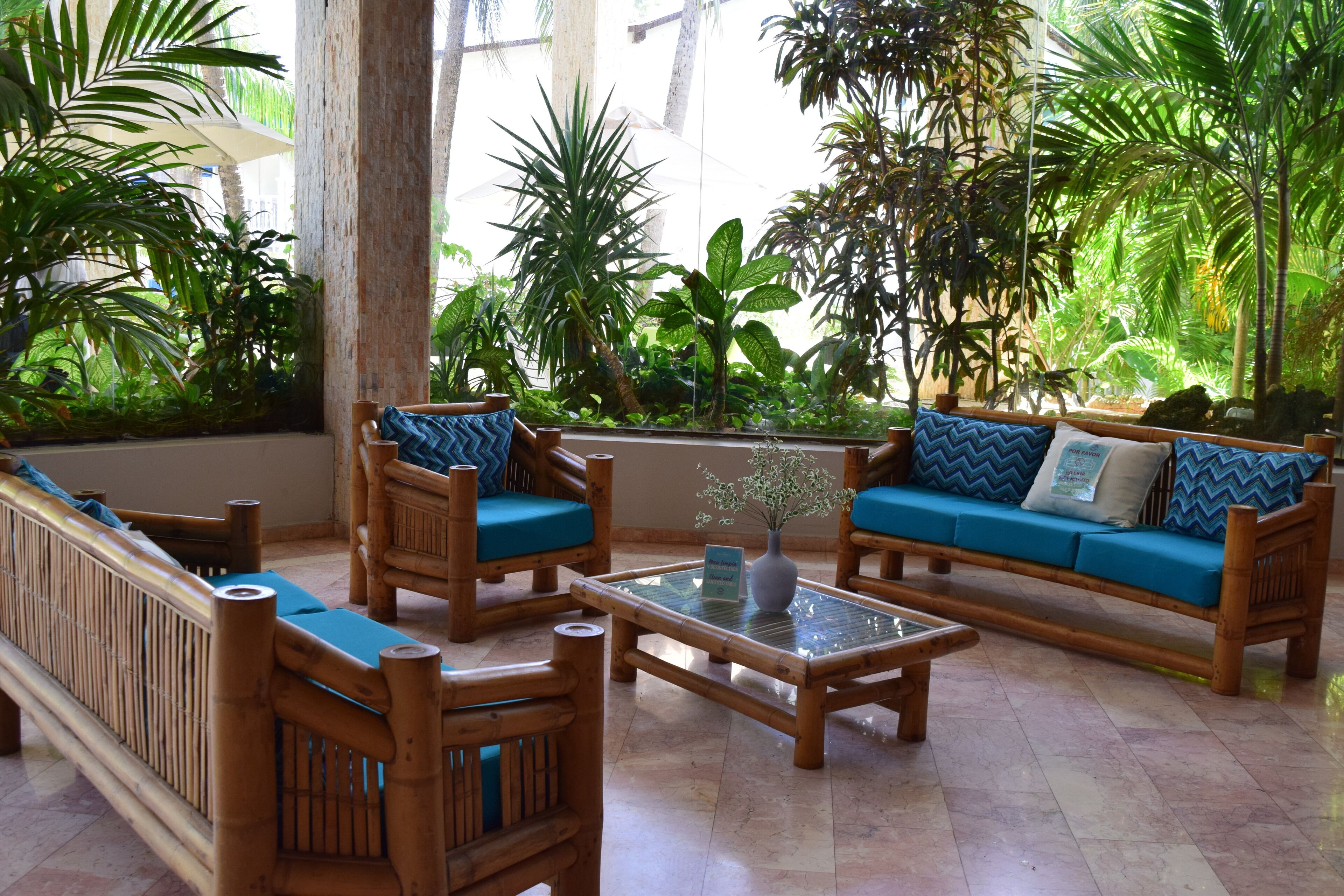 Un área de estar de madera con sofás y sillones tapizados en azul, una mesa de centro de vidrio y un ventanal con vistas a un jardín exuberante.