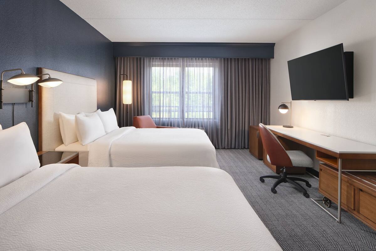 Ein Hotelzimmer mit zwei Betten, einem Schreibtisch, einem Stuhl, einem Fernseher und einem Fenster mit Vorhängen.