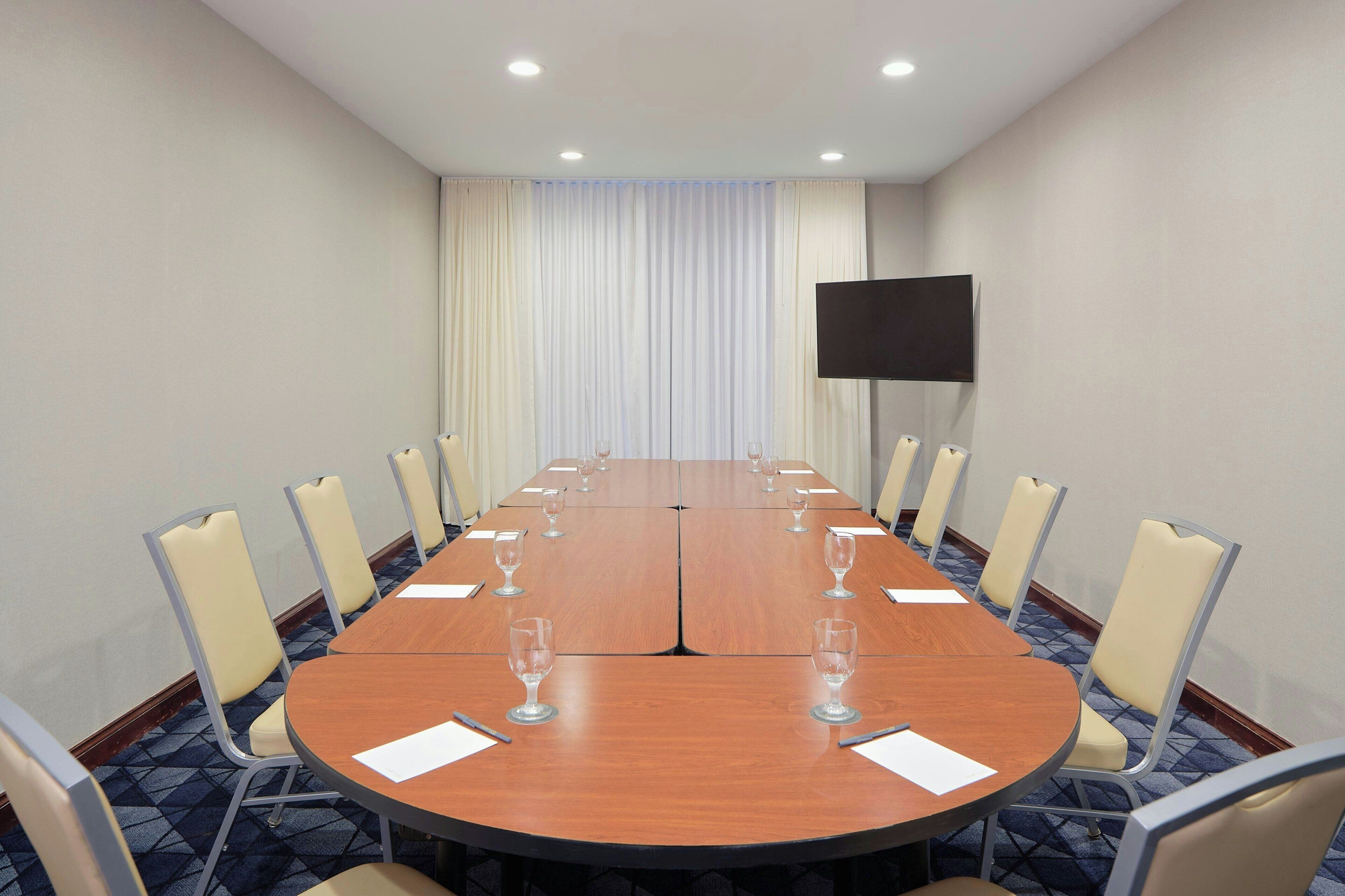 Une salle de conférence avec une longue table, des chaises, un téléviseur à écran plat et une fenêtre avec des rideaux.