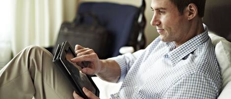 Een man zit op een bed in een hotelkamer en houdt een tablet vast.